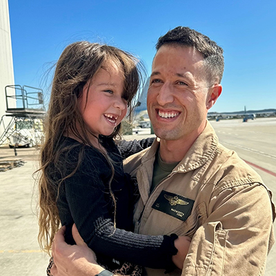 A man in a military battle jacket holds a young girl who is smiling and missing front teeth.