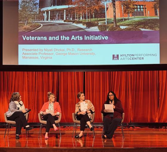 Four women sit on stage before a powerpoint titled "Veterans and the Arts Initiative"